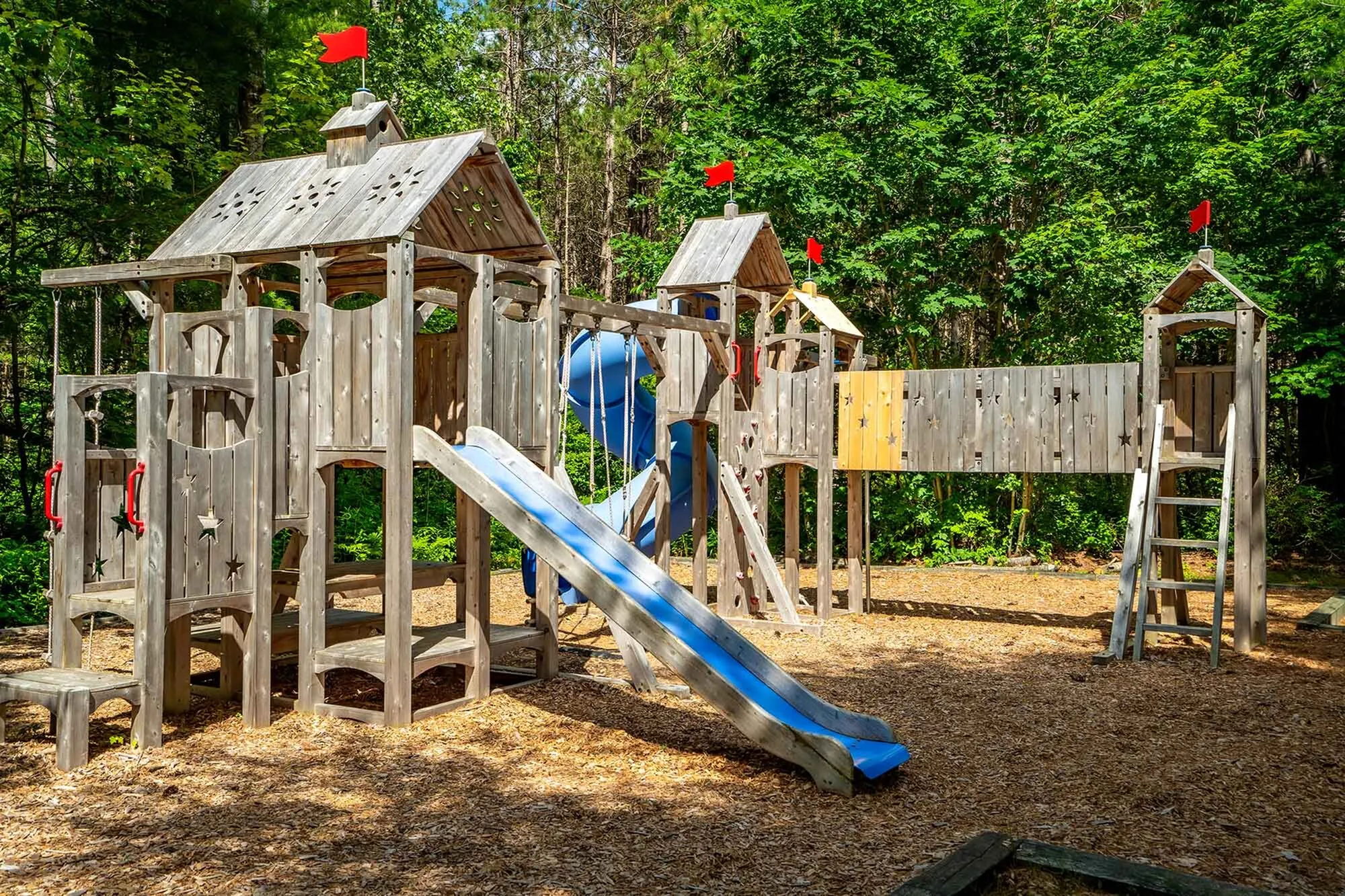Enchanting wooden playground in a lush forest setting features slides and climbing towers with playful red flags.