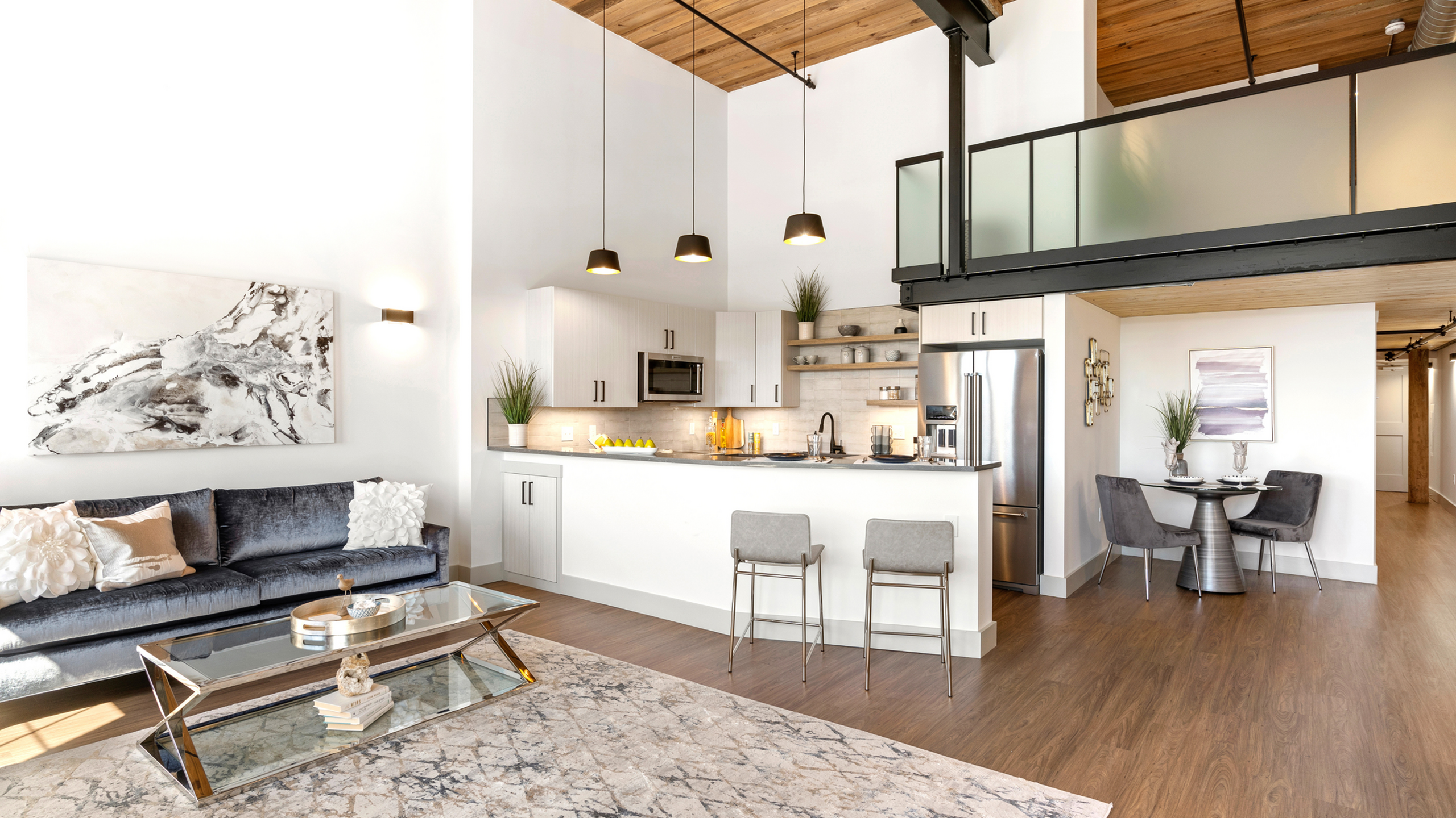 Riverwalk Lofts | A modern loft apartment with an open-concept living space featuring high ceilings and natural wood finishes. The kitchen includes stainless steel appliances, pendant lighting, and a breakfast bar with seating. A cozy livin