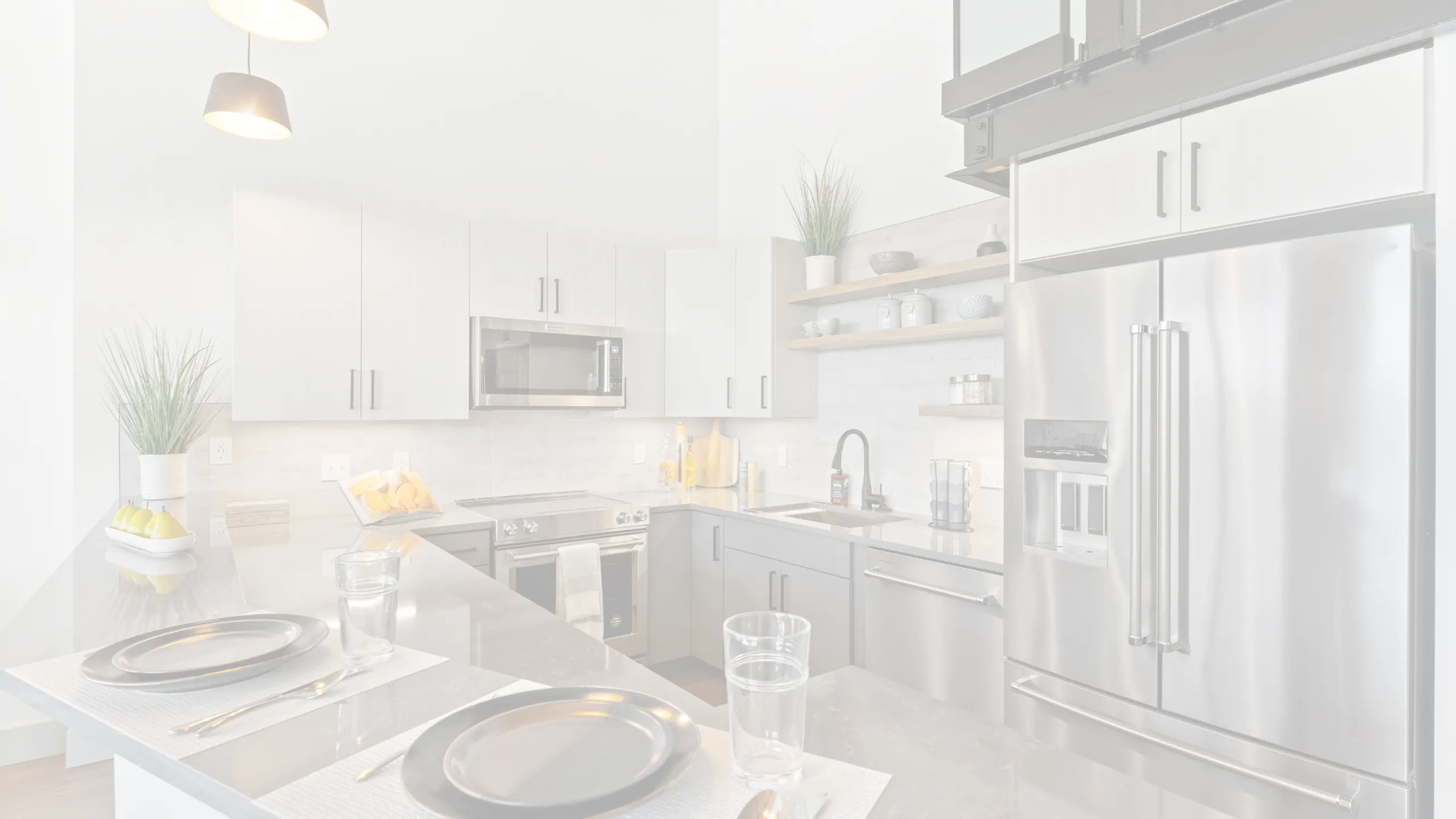 Riverwalk Lofts | Modern kitchen with stainless steel appliances, including a fridge and oven. White cabinets, open shelving with decor, and a dark island with two place settings. Pendant lights hang above the island, and potted plants add
