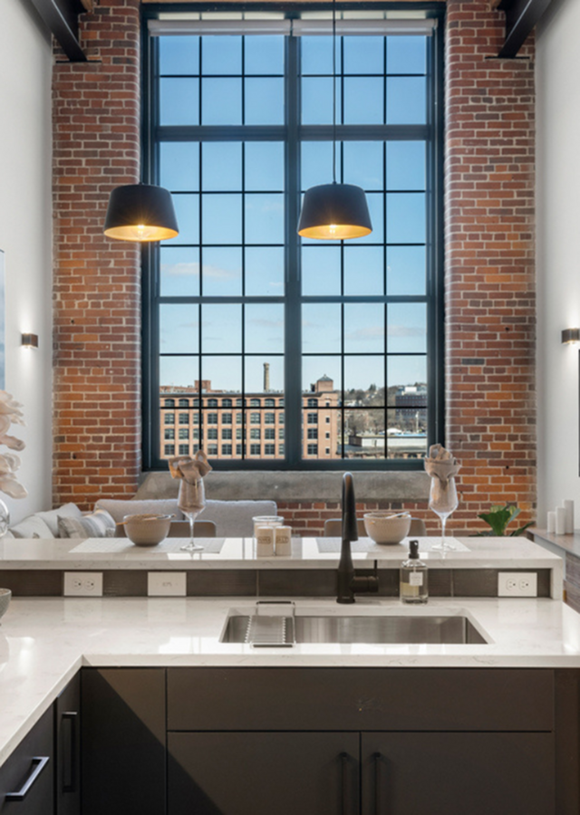 Riverwalk Lofts | A modern kitchen with a marble countertop and a black faucet. Large industrial-style windows reveal a cityscape with brick buildings. Two pendant lights hang above the counter, and decorative items adorn the space. | Studi