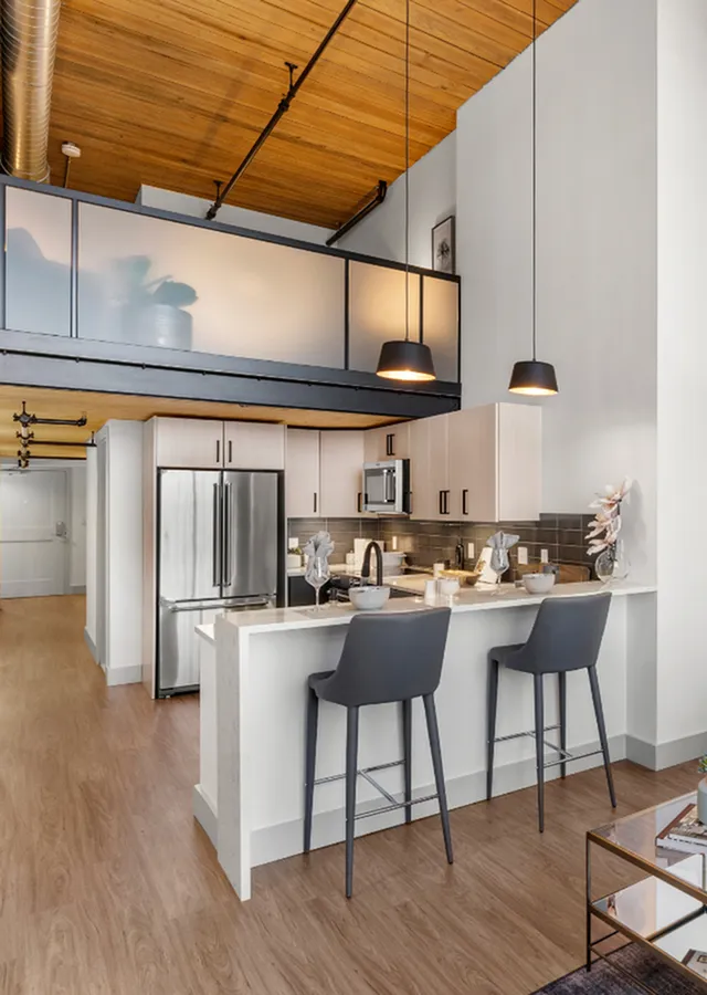 riverwalk lofts | Modern kitchen with high ceilings and wooden floors, featuring white cabinetry, a stainless steel refrigerator, and a breakfast bar with two gray stools. The space includes pendant lighting and a loft area above. | studio