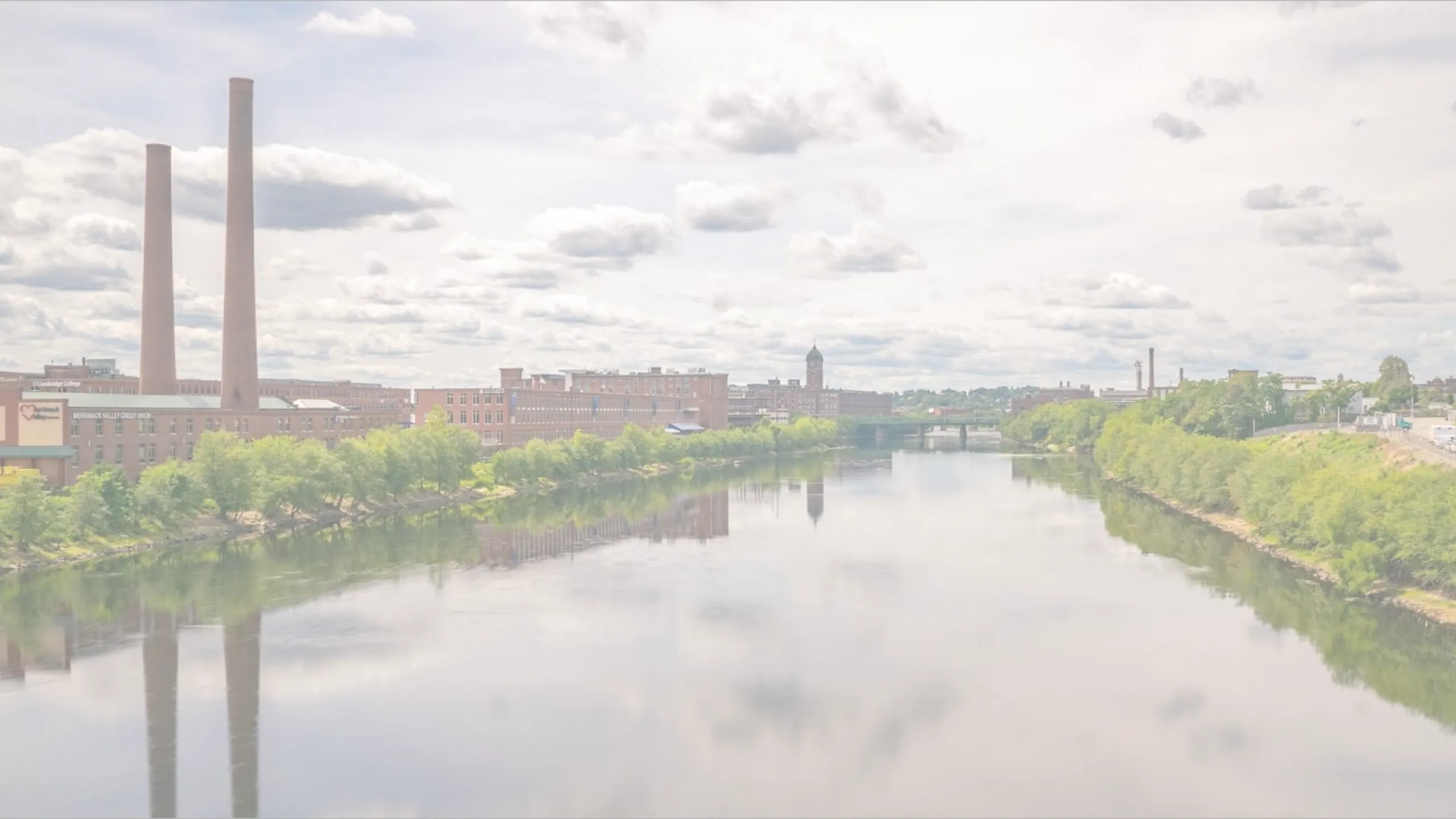 Riverwalk Lofts | A river flows between two banks lined with industrial buildings and tall chimneys, reflecting the cloudy sky. Lush green trees border the water, with a distant bridge visible in the background. | Apartments for rent lawren
