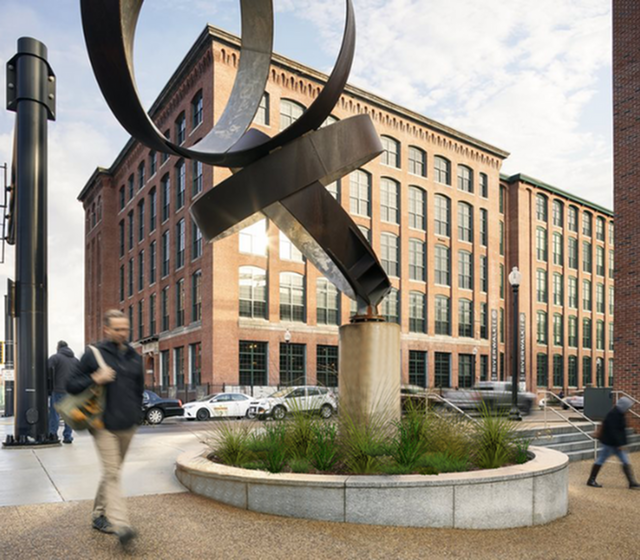 riverwalk lofts | A street scene with a large abstract metal sculpture in front of a brick building. People walk by on the sidewalk, and a few cars are parked along the street. The sky is partly cloudy. | apartments for rent in lawrence ma