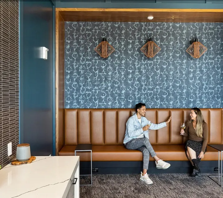 Riverwalk Lofts | A modern lounge area with two people sitting on a brown leather bench, raising drinks while smiling at each other. The wall features a patterned design with three geometric art pieces. A white marble counter is visible in