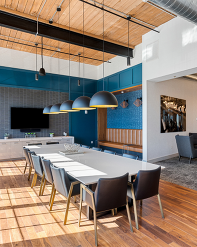riverwalk lofts | Modern meeting room with a long white table, black chairs, and hanging pendant lights. The room features a wood ceiling, blue accent wall, mounted TV, and decorative wall art. Natural light streams in through large windows
