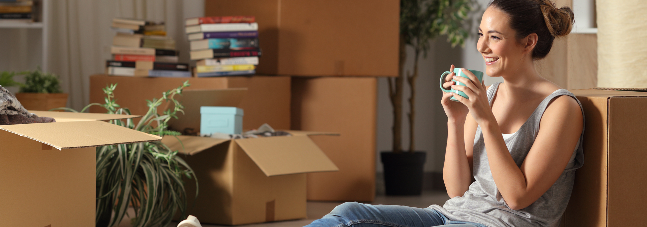 Smiling woman enjoying a drink surrounded by moving boxes and plants, symbolizing a fresh start in a new home.