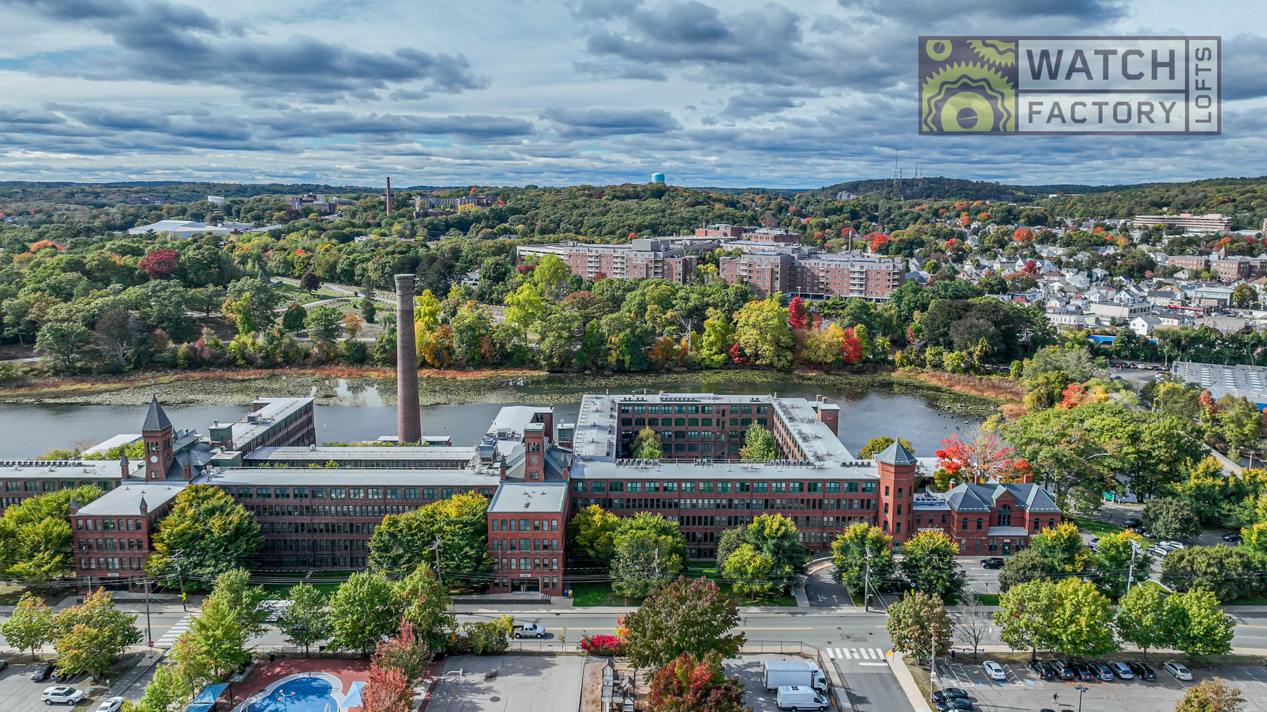 Aerial view of Watch Factory Lofts surrounded by scenic river and lush trees under a cloudy sky.