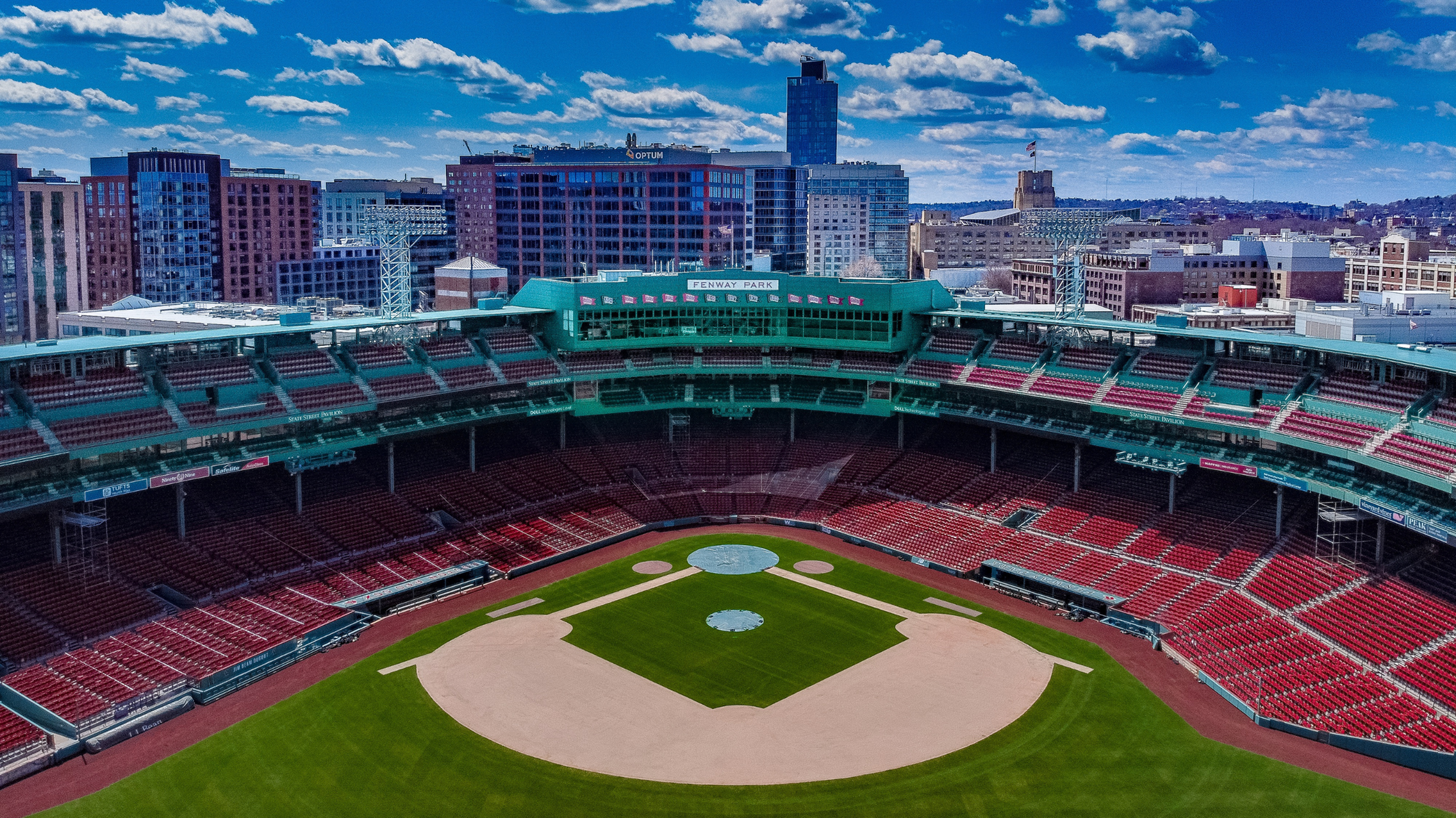 Aerial view of a Fenway baseball stadium with a lush green field, surrounded by red seats and cityscape in the background.