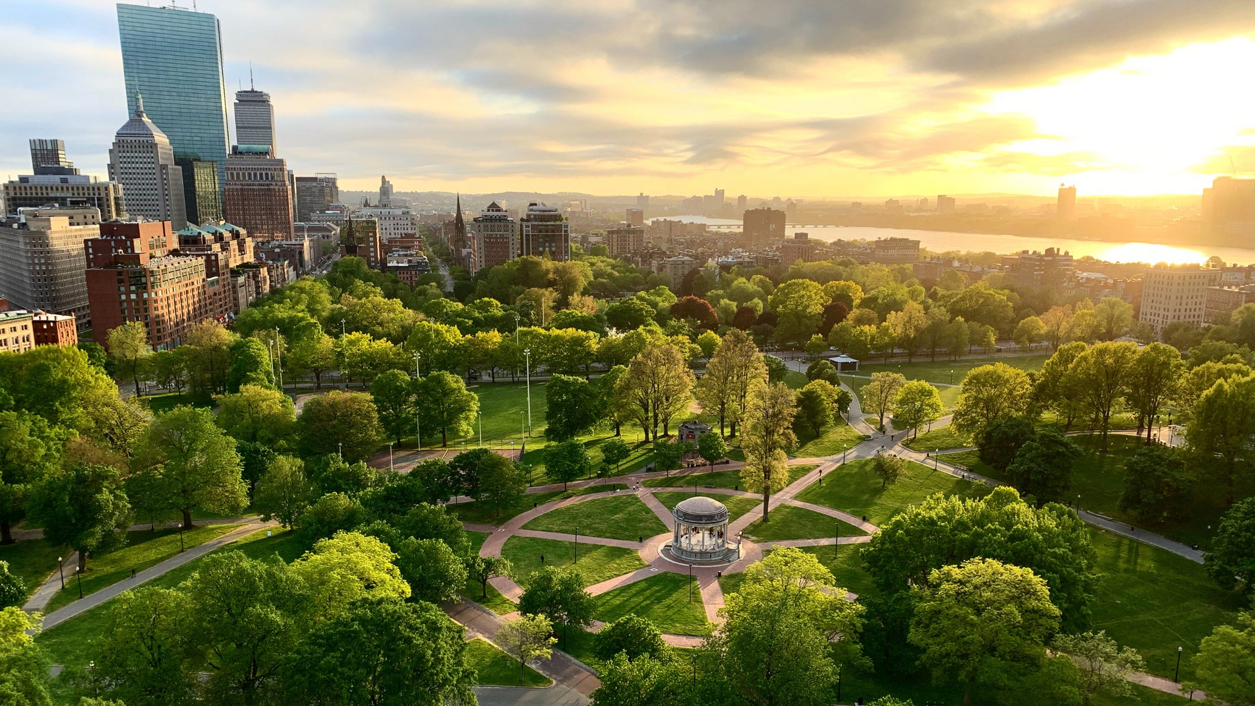 Aerial view of a vibrant Boston city park surrounded by lush green trees with a skyline of tall buildings at sunset.