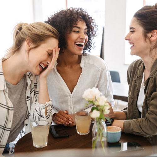 The James Apartments | Three friends enjoying laughter and drinks in a cozy, sunlit caf&eacute; setting. | Haverhill Apartments with Balcony