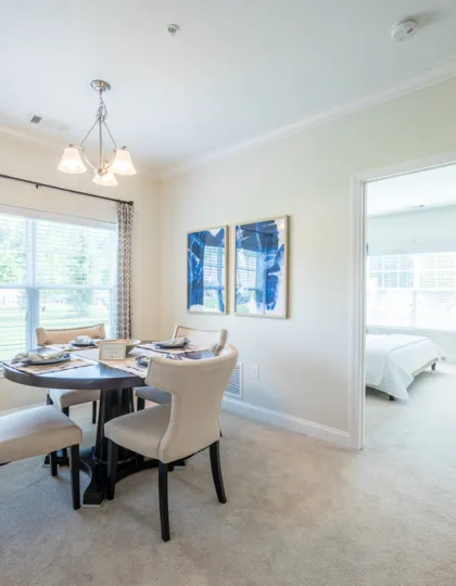 Bright dining area with elegant furniture, natural light, and soothing wall art leading to a cozy bedroom.