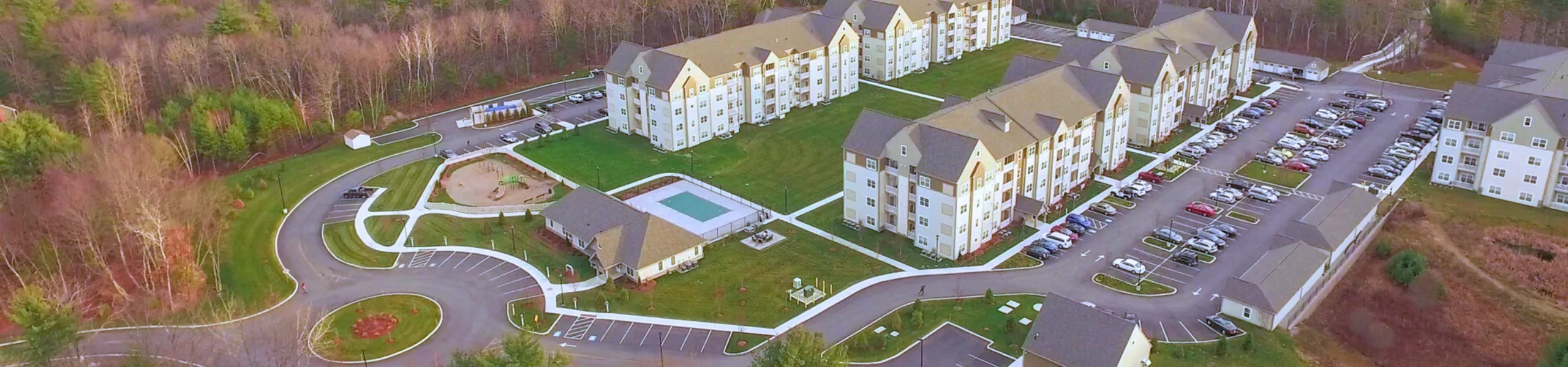 Scenic aerial view of modern apartment complex surrounded by lush greenery, featuring ample parking and inviting amenities like a playground and swimming pool.