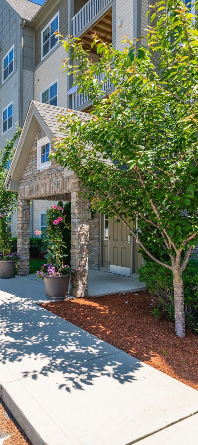 Charming apartment entrance with elegant stonework, vibrant potted flowers, and lush green landscaping under a sunny blue sky.