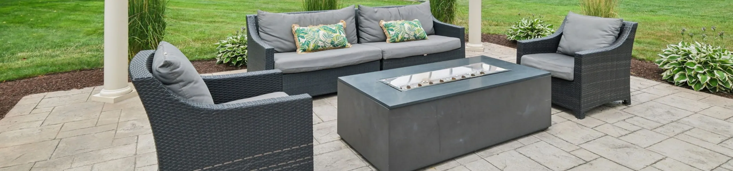 Princeton Westford Apartments | Modern outdoor patio at Princeton Westford Apartments featuring a white pergola, gray wicker furniture, a community patio with firepit, decorative plants, and views of green trees and parked cars in the backg
