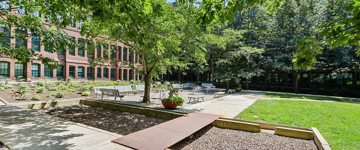 Charming garden courtyard with lush trees, seating area, and vibrant red brick building backdrop.