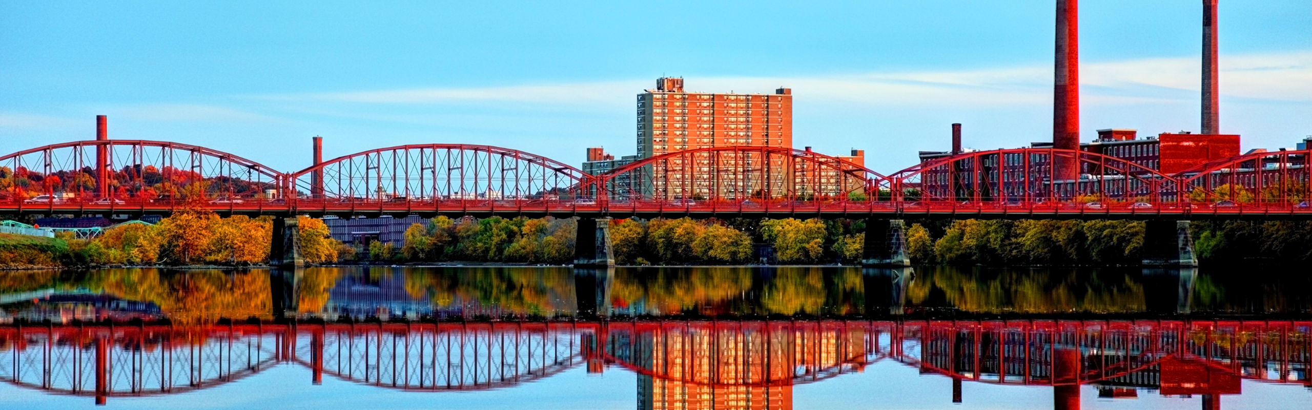 Vibrant Lowell MA featuring a stunning red bridge reflecting on calm waters, with modern high-rise buildings and lush autumnal trees under a clear blue sky.