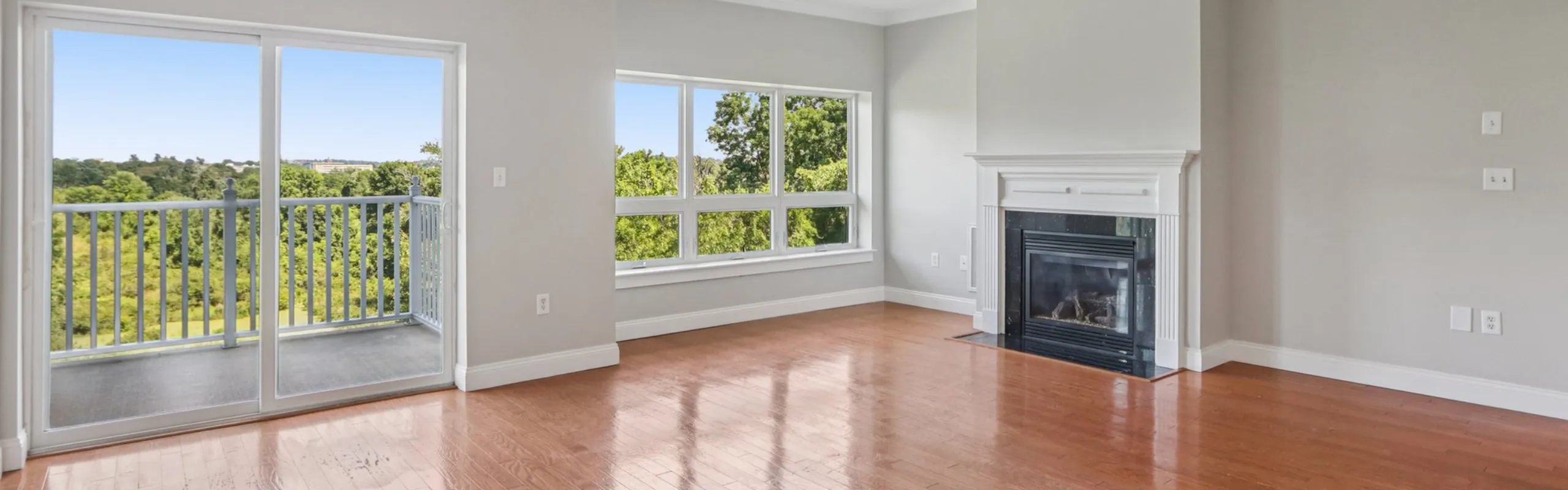 Bright living room featuring hardwood floors, a modern fireplace, and large windows with stunning green views and balcony access.