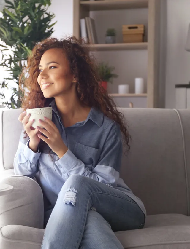 Grandview Apartments | Cozy and inviting living space with a relaxed ambiance, featuring a woman enjoying a warm beverage on a stylish gray sofa. | Pet Friendly apartments in lowell ma