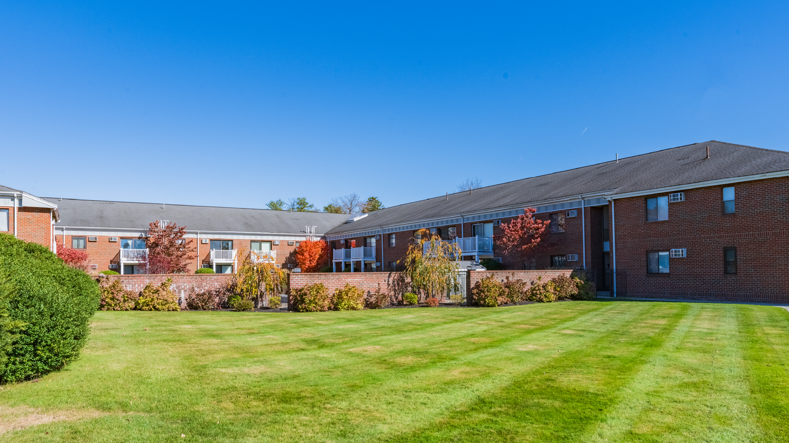 Charming brick apartment complex with private balconies, lush green lawn, and vibrant autumn foliage under a clear blue sky.