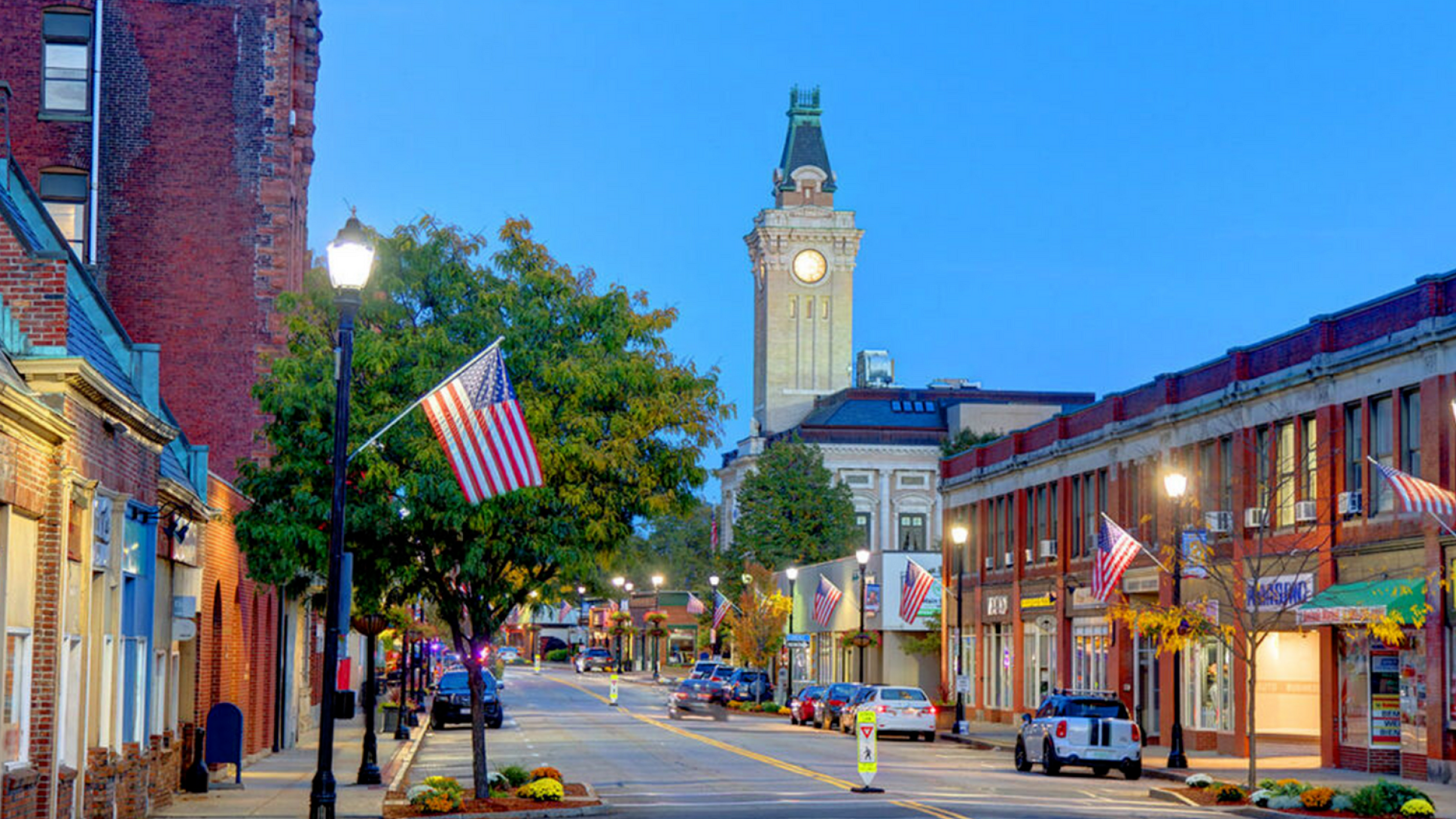 Charming small-town street scene at dusk with historic clock tower, vibrant storefronts, and American flags lining the sidewalk.