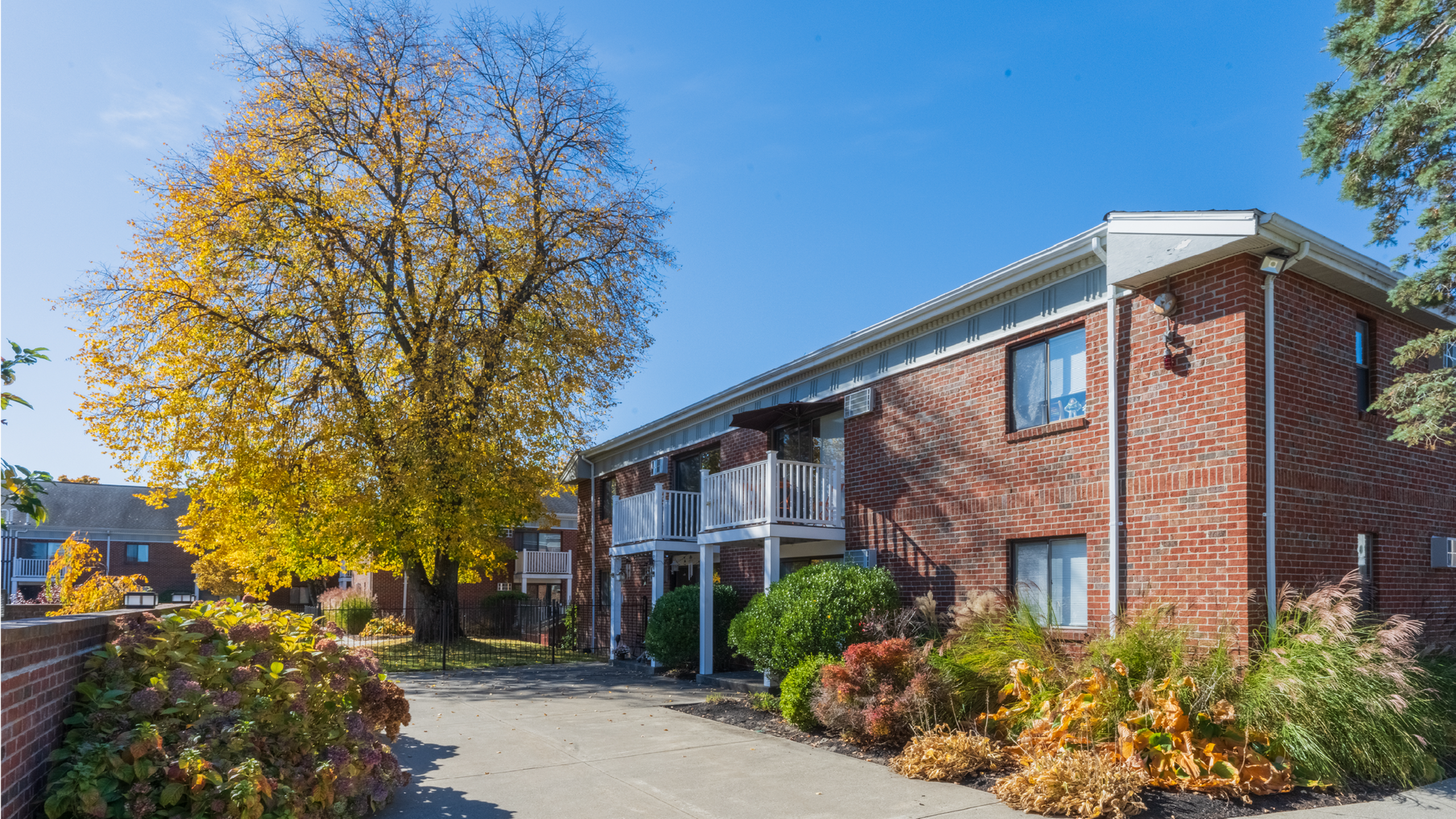 Charming brick apartment building with inviting balconies, surrounded by lush landscaping and vibrant autumn trees, set against a clear blue sky.