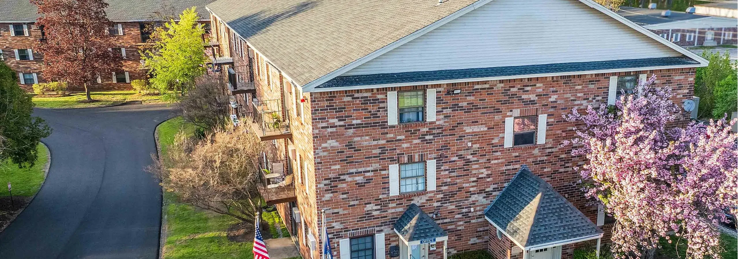 Charming brick apartment building surrounded by lush greenery and vibrant blooming trees, offering a welcoming and tranquil living environment.