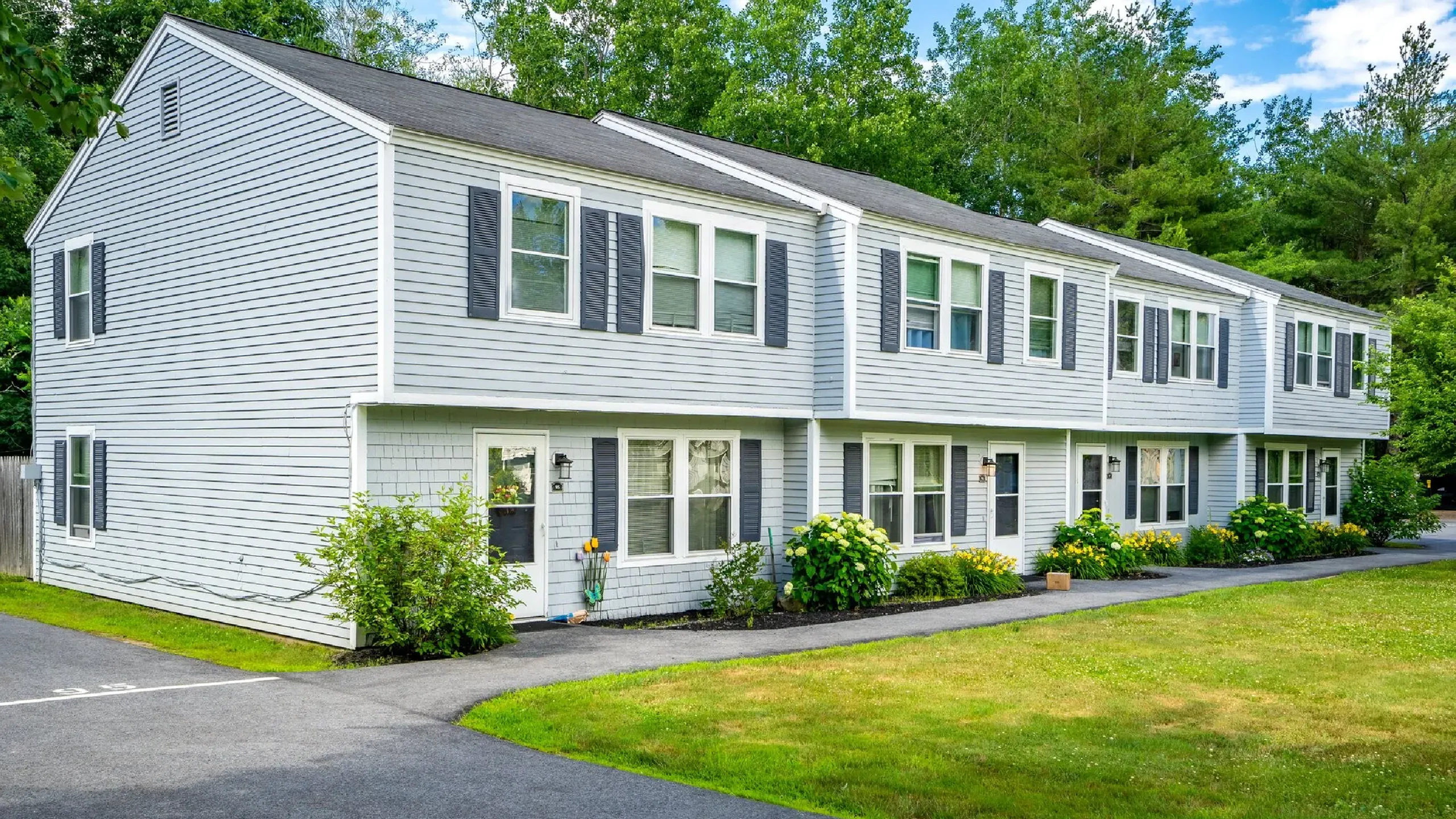 Charming duplex surrounded by lush greenery, featuring a well-maintained exterior with classic shutters and a vibrant garden.