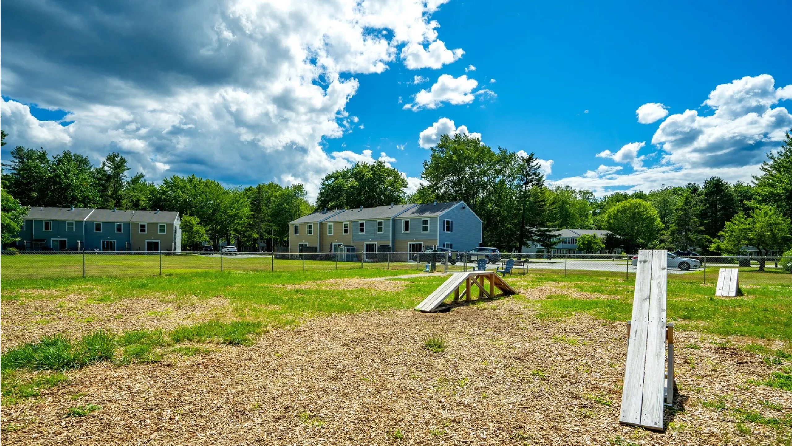 Expansive dog park with agility ramps, surrounded by charming suburban townhouses and lush greenery under a vibrant blue sky.