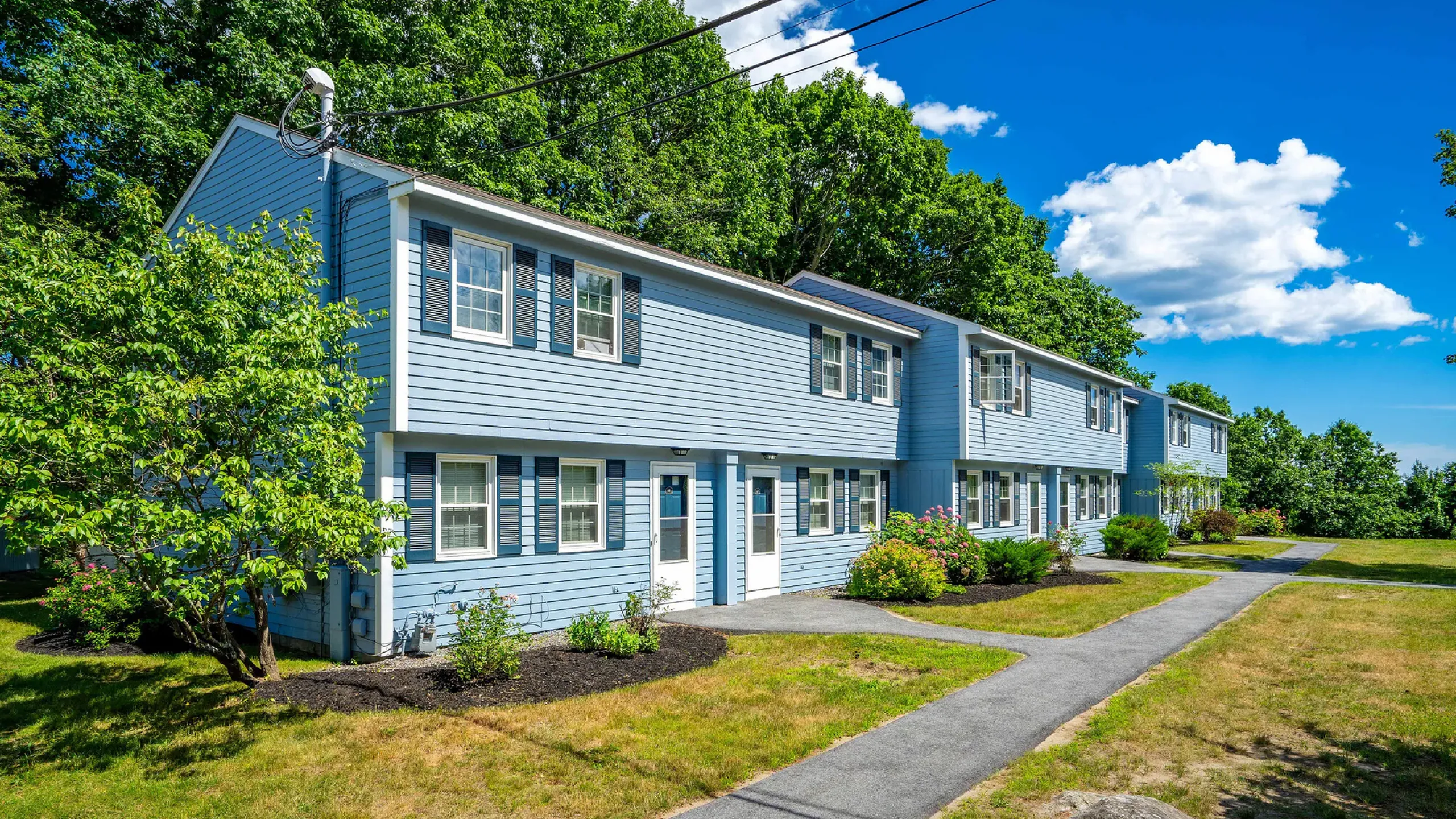 Charming blue townhouse complex surrounded by lush greenery and bright flowers under a vibrant blue sky.