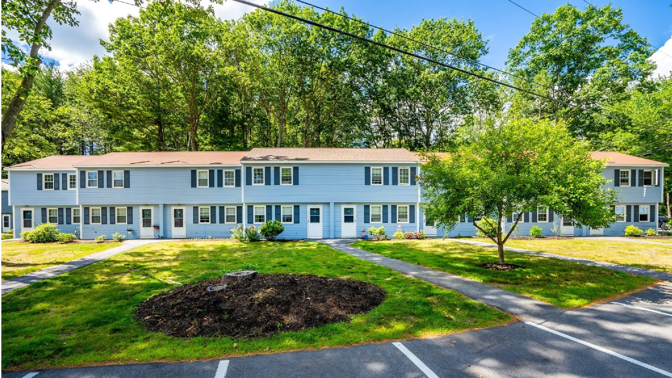 Charming blue apartment complex surrounded by lush greenery, vibrant landscaping, and clear skies, offering a serene and inviting living environment.
