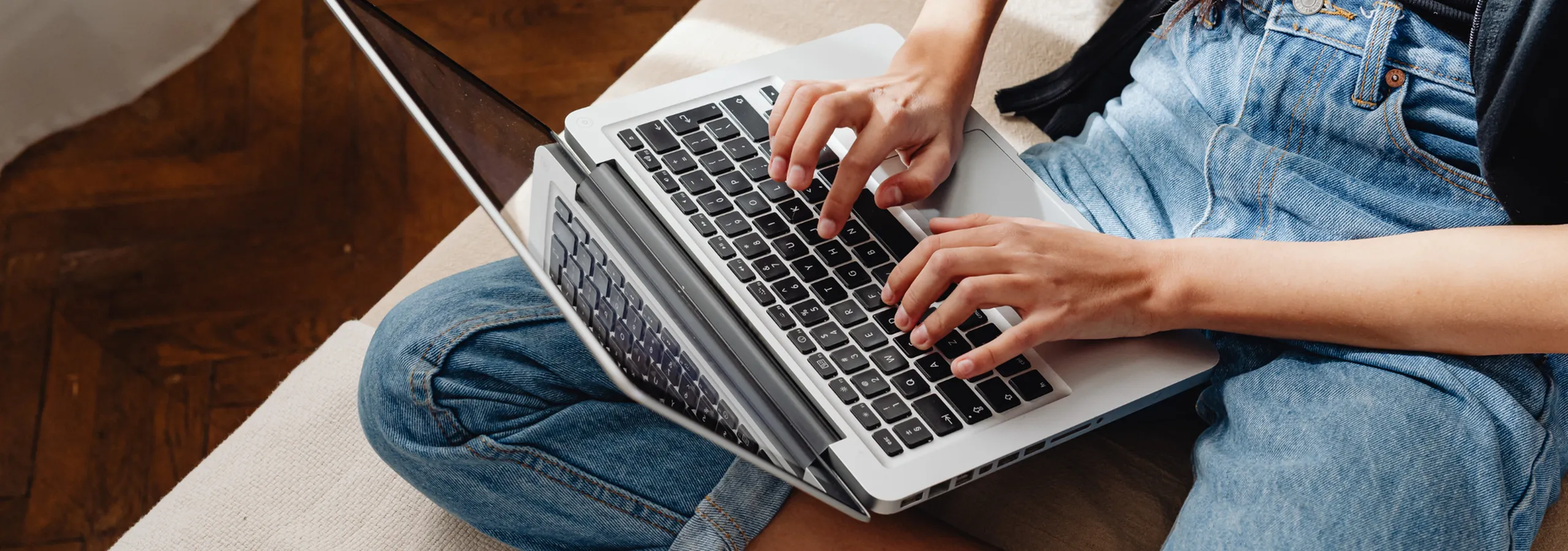Focused individual typing on a sleek laptop, sitting comfortably on a cozy beige sofa, highlighted by a vibrant wooden floor.