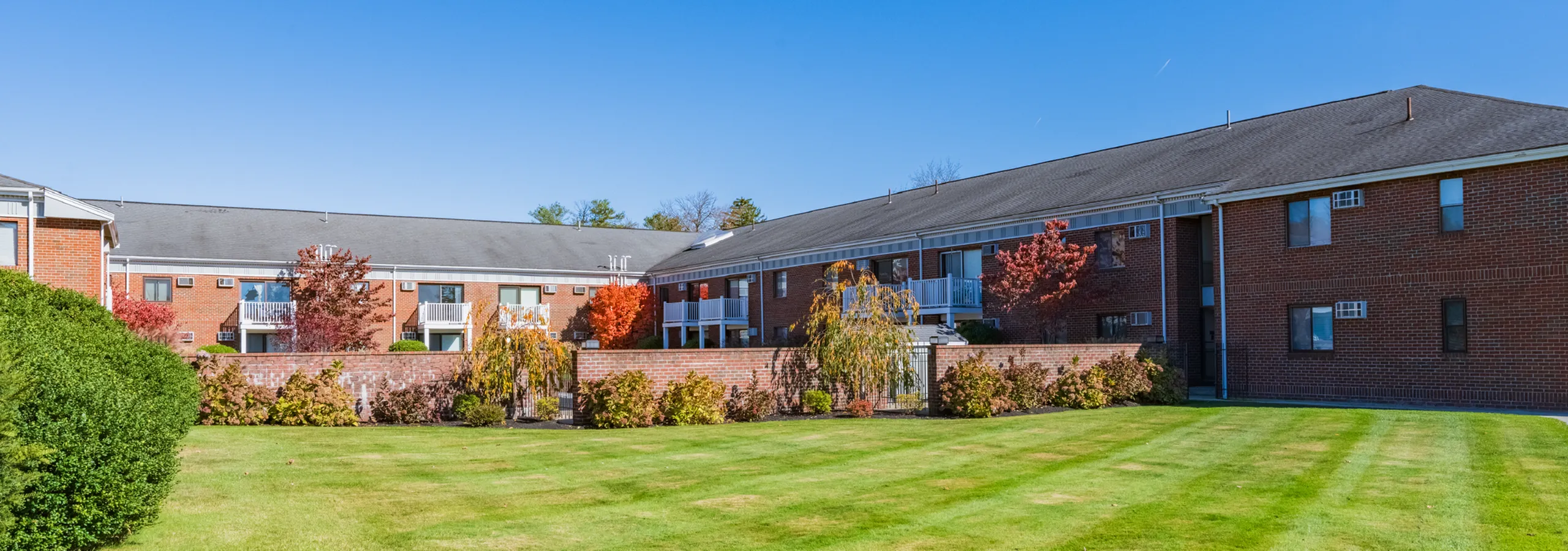 Charming apartment complex with red brick exterior, lush green lawns, and vibrant autumn foliage under a clear blue sky.
