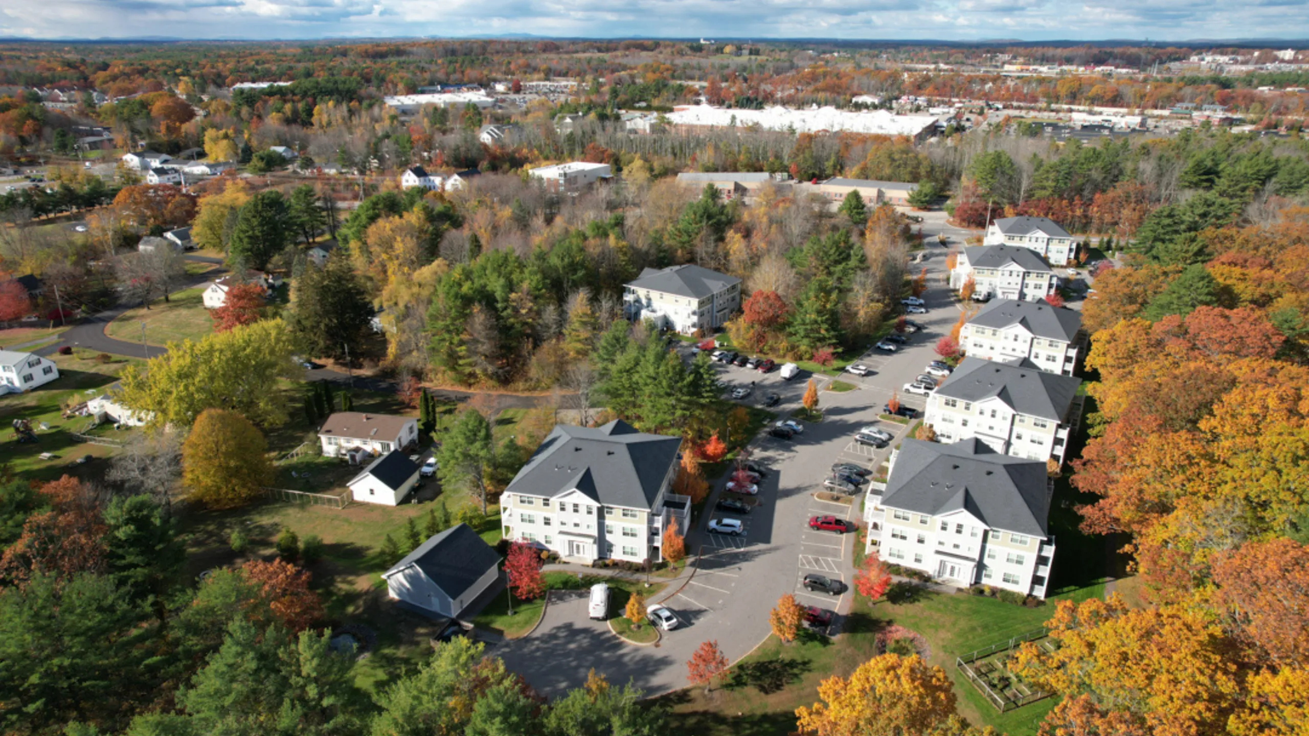 Charming aerial view of luxurious apartments nestled amidst vibrant autumn foliage, offering a serene wooded landscape and ample parking.