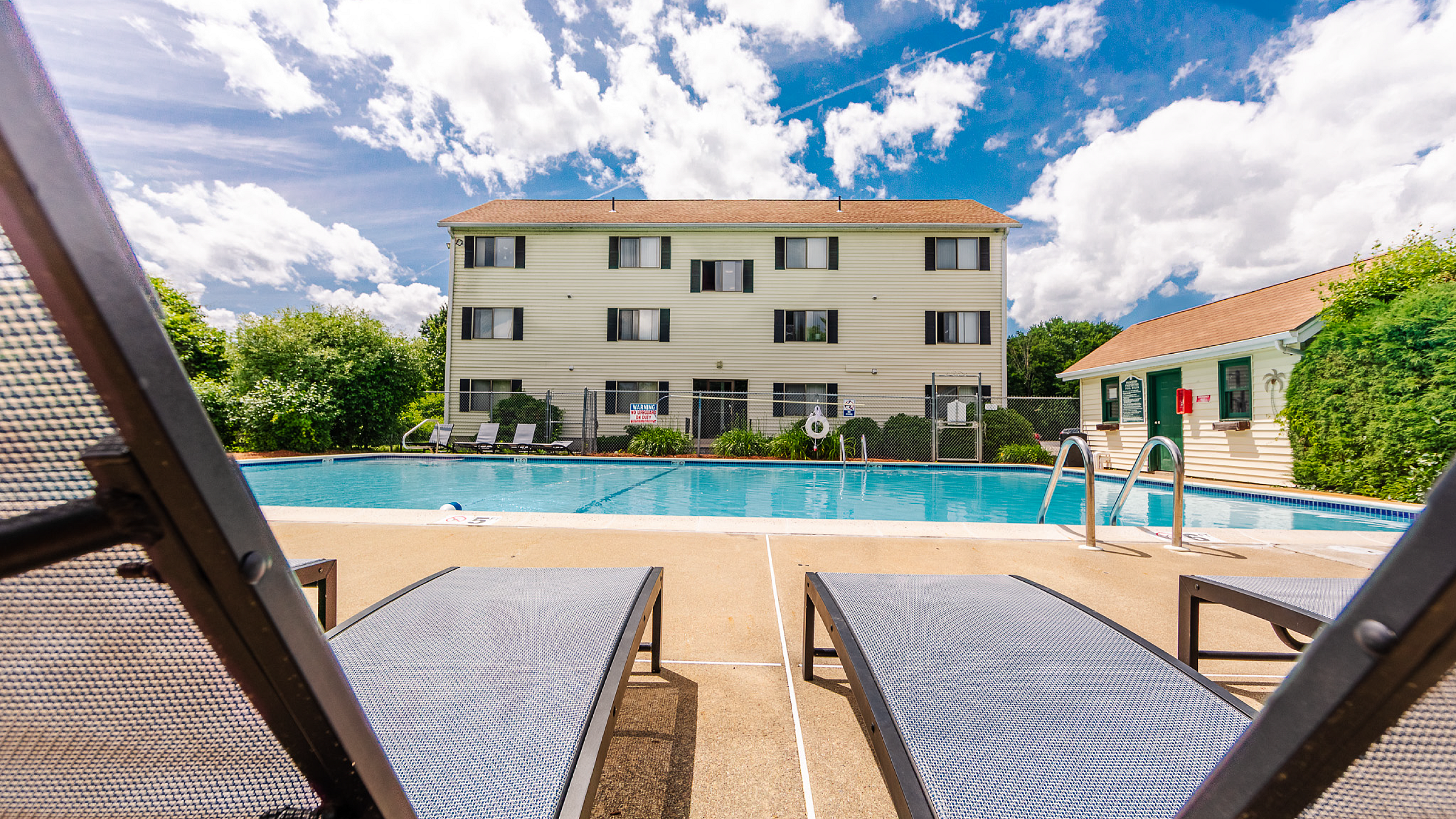 Inviting apartment complex with a refreshing swimming pool, surrounded by lush greenery and sun loungers under a vibrant blue sky.