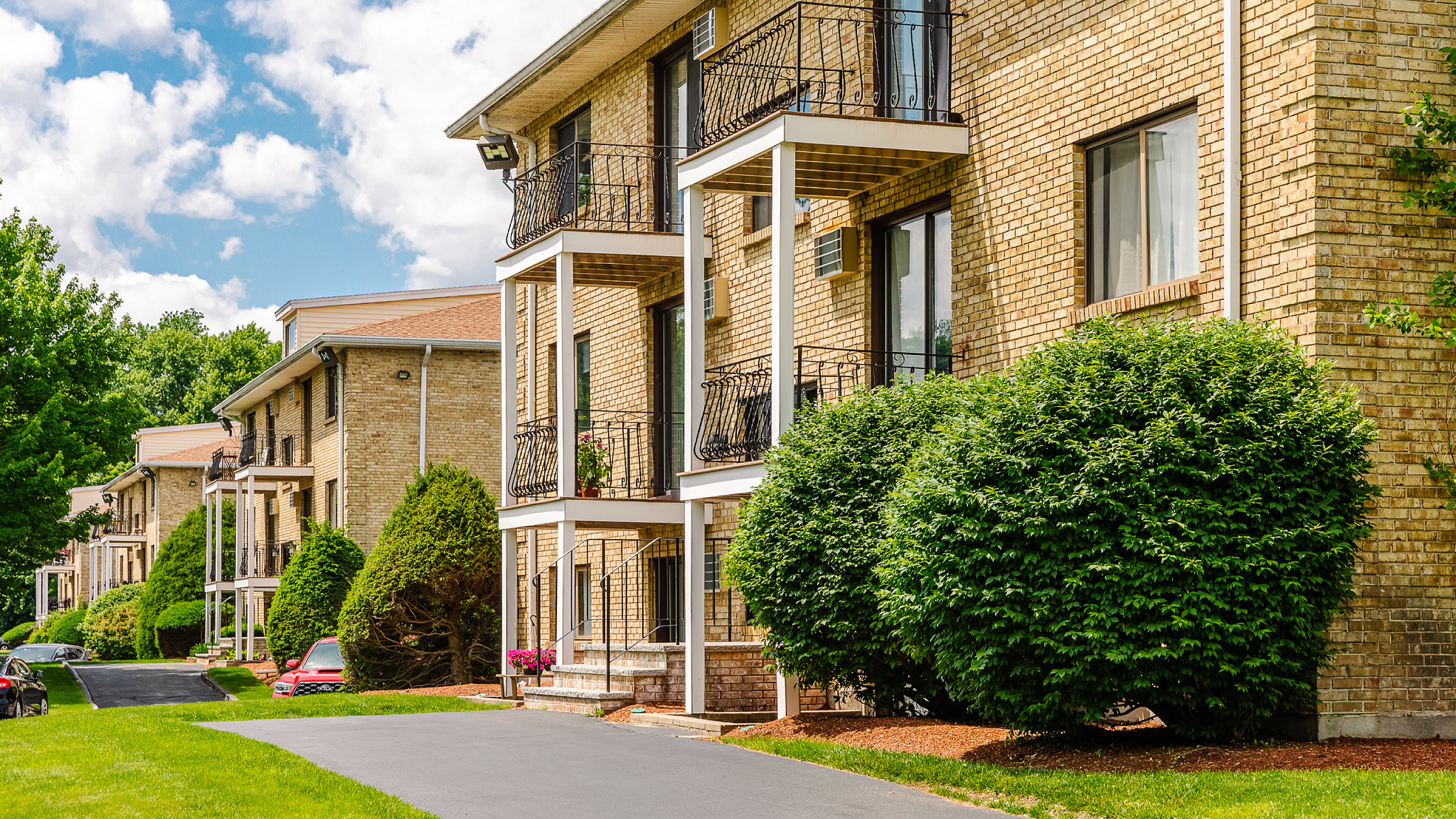 Charming apartment complex with classic brick exterior, private balconies, and lush green landscaping on a sunny day.