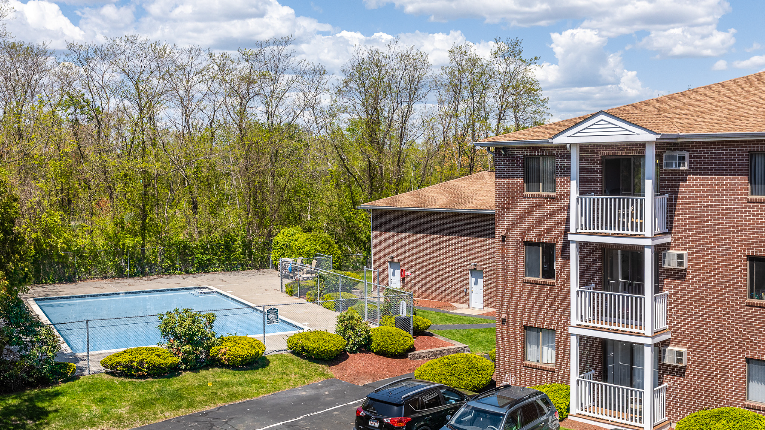 Charming brick apartment building with private balconies next to a refreshing outdoor pool, surrounded by lush greenery under a bright blue sky.