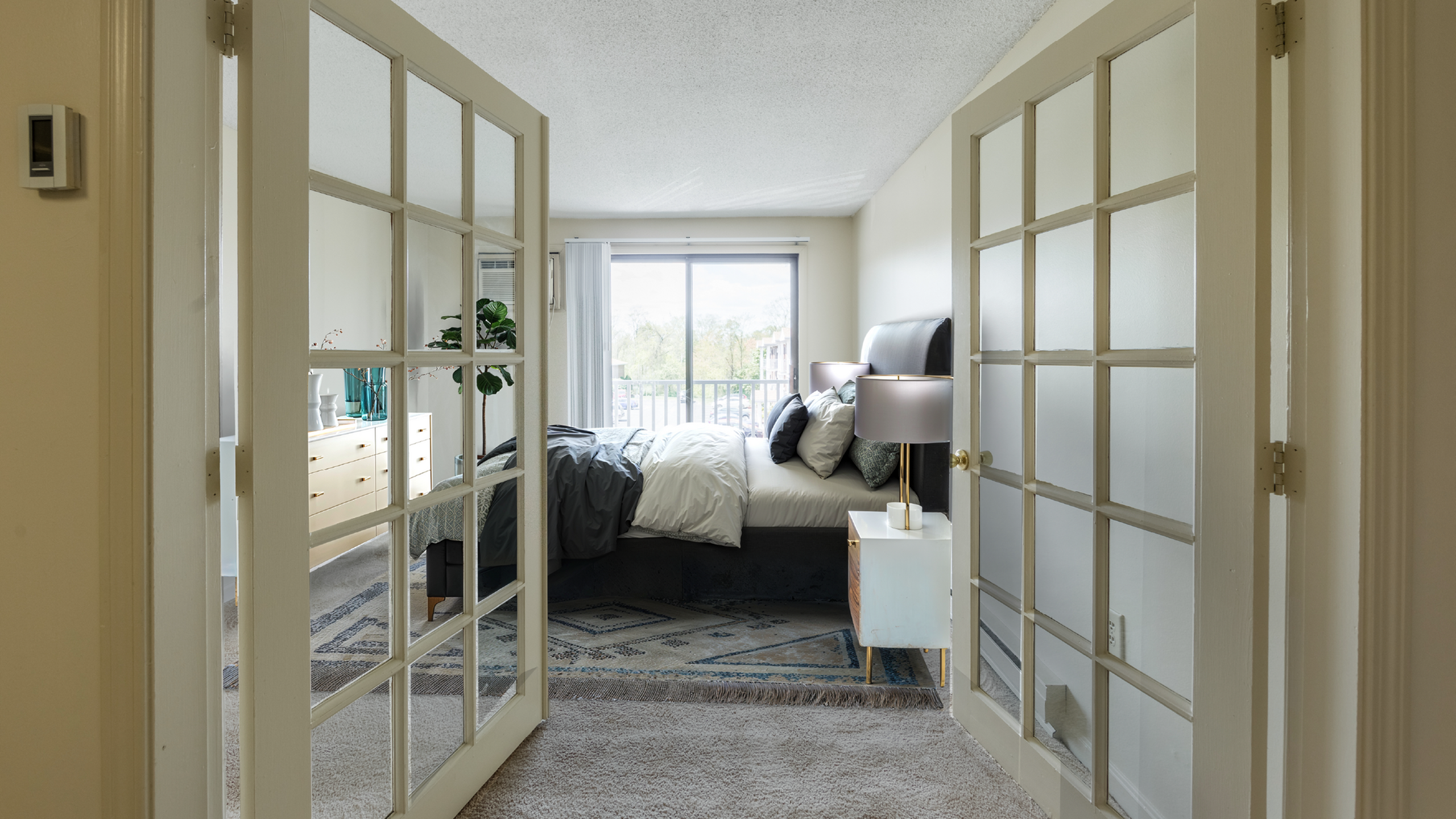 Elegant bedroom with glass French doors, plush bedding, and a bright window view, creating a serene and inviting atmosphere.
