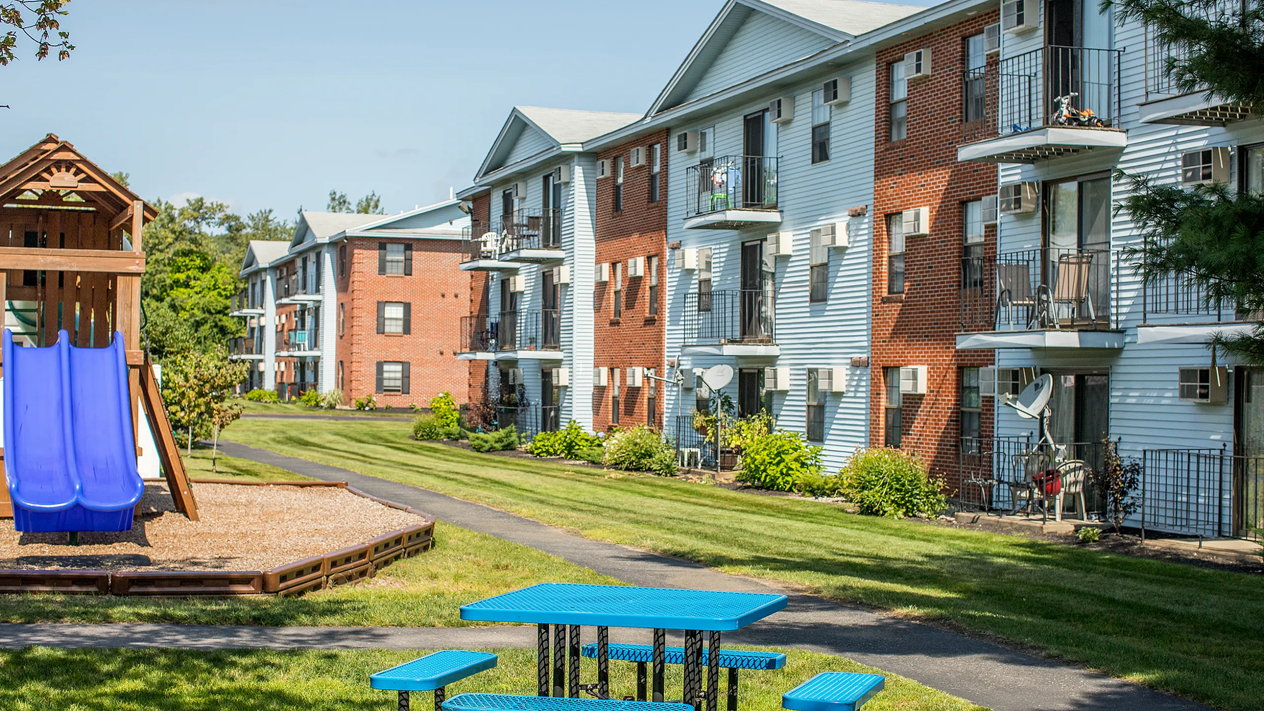Charming apartment buildings with balconies, surrounded by lush green lawns and a friendly playground area, ideal for families.
