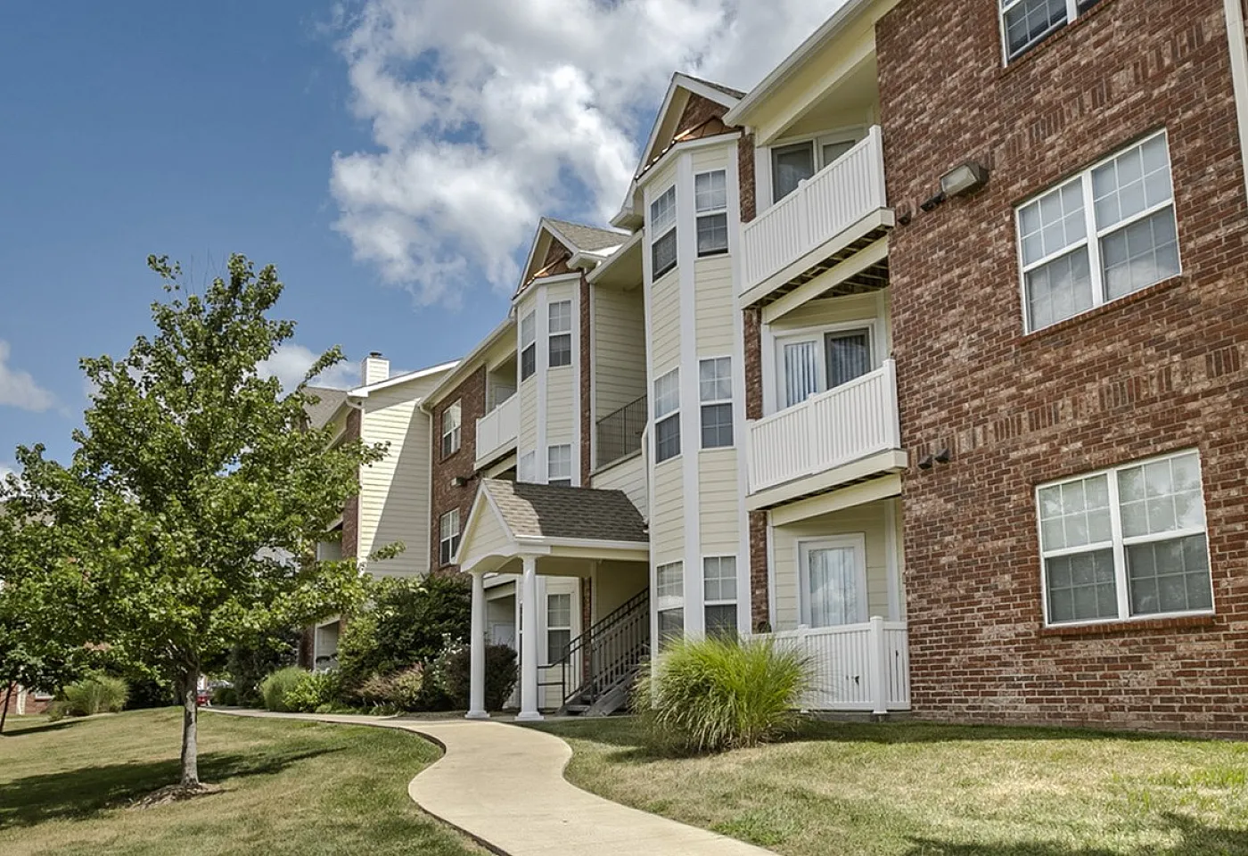 Charming brick apartment building surrounded by lush green lawns and vibrant trees under a bright blue sky.