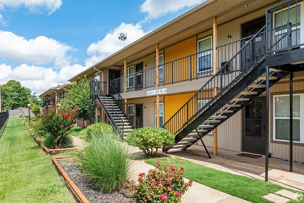 Charming two-story apartment complex with vibrant yellow accents, lush landscaping, and spacious outdoor walkways under a clear blue sky.