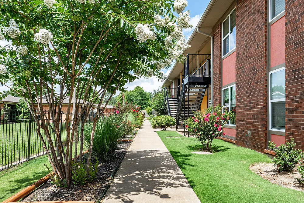 Charming apartment exterior with vibrant landscaping, blooming flowers, and a welcoming walkway leading to the building entrance.