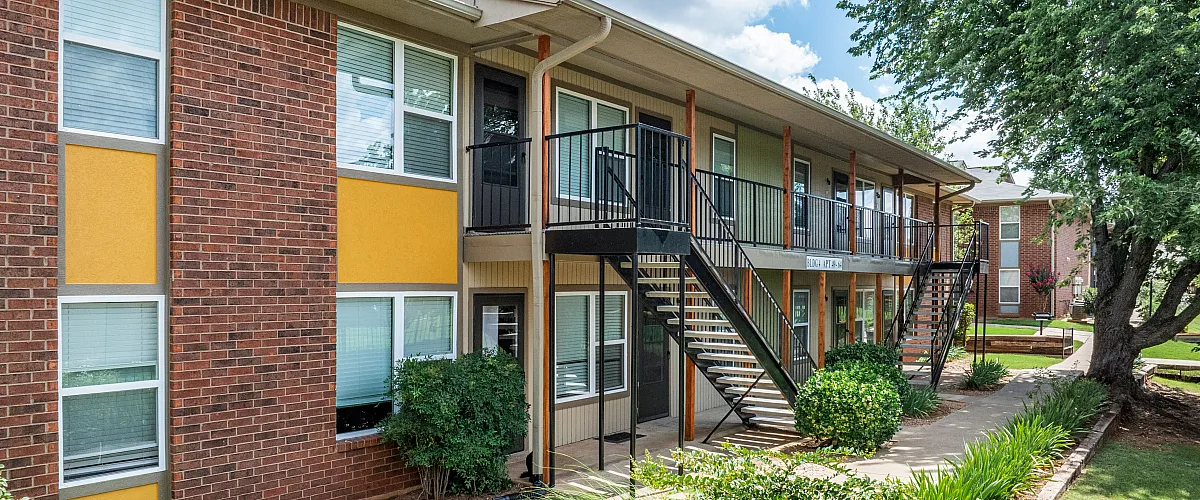 Charming apartment complex with vibrant brick and yellow facade, lush landscaping, and inviting stairway access.
