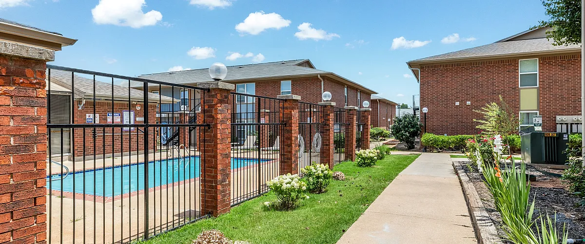 Inviting apartment courtyard with a sparkling fenced pool, vibrant red brick buildings, lush greenery, and a well-maintained pathway under a clear blue sky.