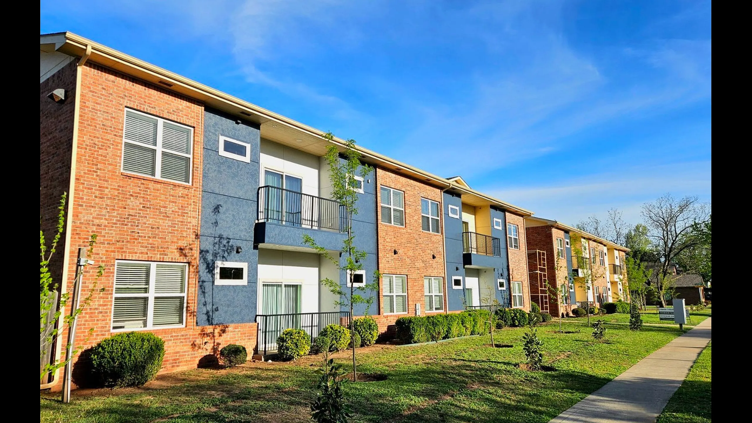Charming two-story apartment building with vibrant red brick and modern blue accents, nestled in a serene landscaped setting under a clear blue sky.