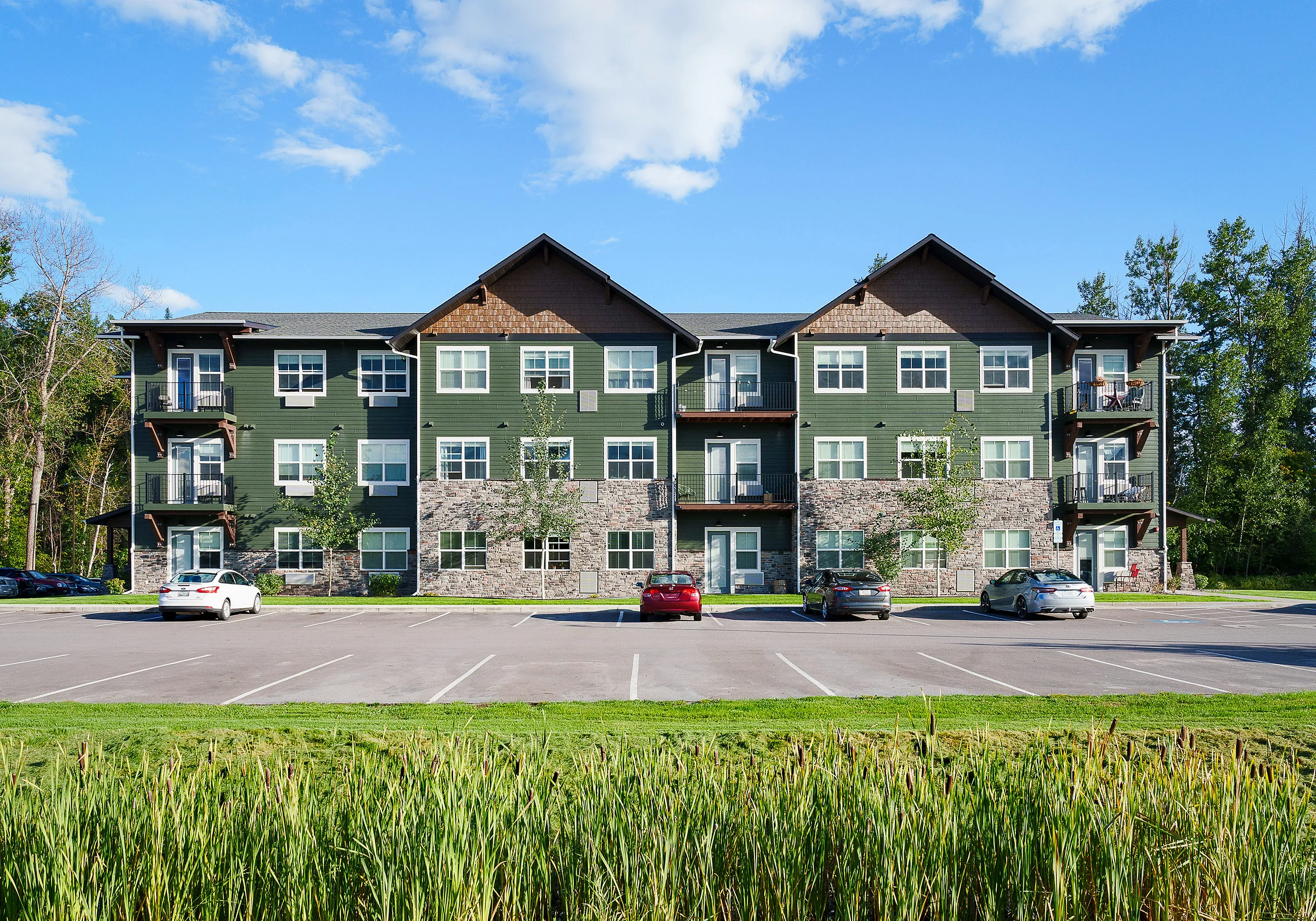 Elegant three-story apartment building with green siding, stone accents, and balconies amidst a scenic landscape.