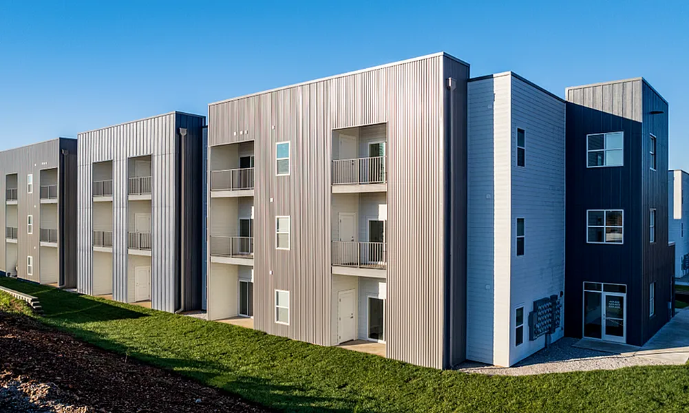 Modern apartment complex with sleek metal facade, spacious balconies, and sunlit green surroundings.