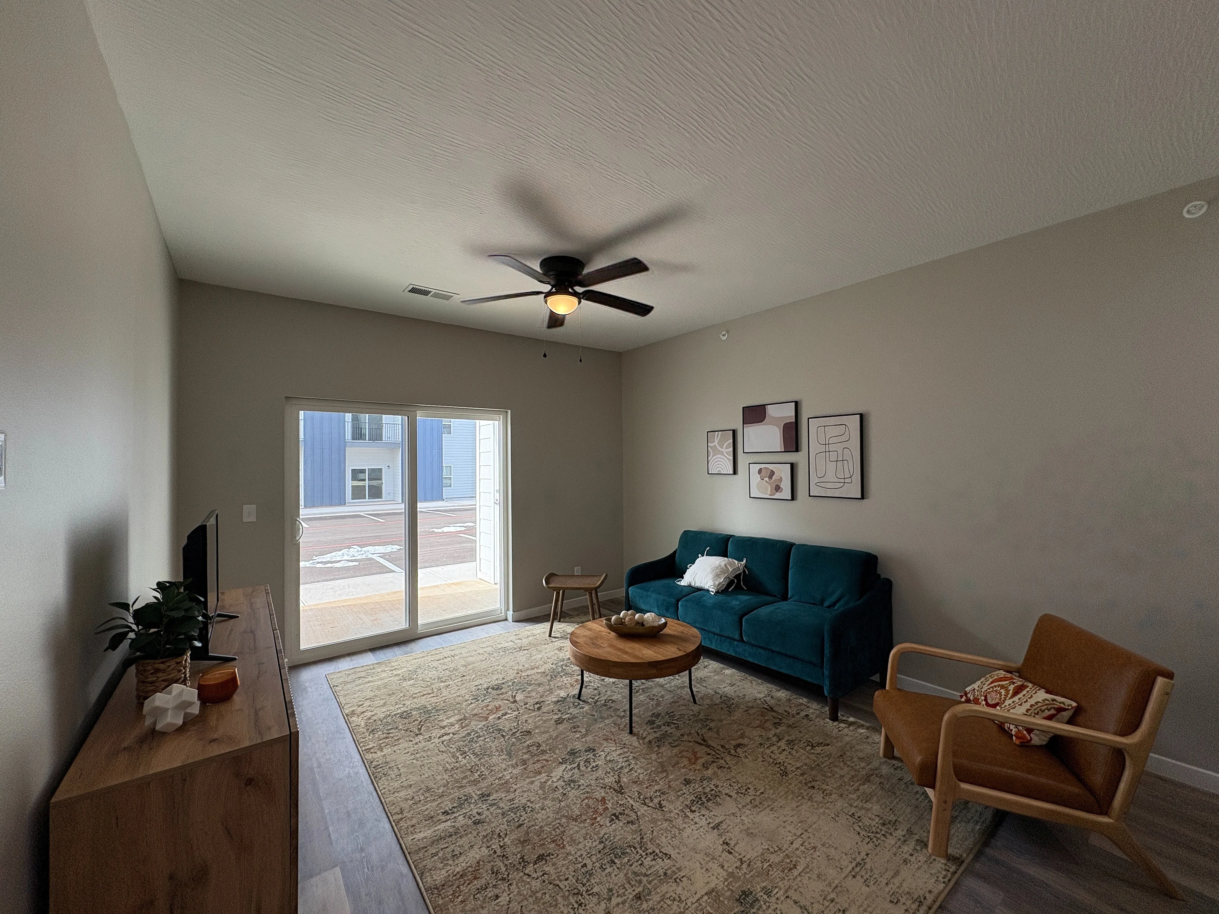 Inviting living room with modern furnishings, featuring a teal sofa, stylish armchair, and wooden accents. Natural light flows through large glass doors, enhancing the cozy ambiance.