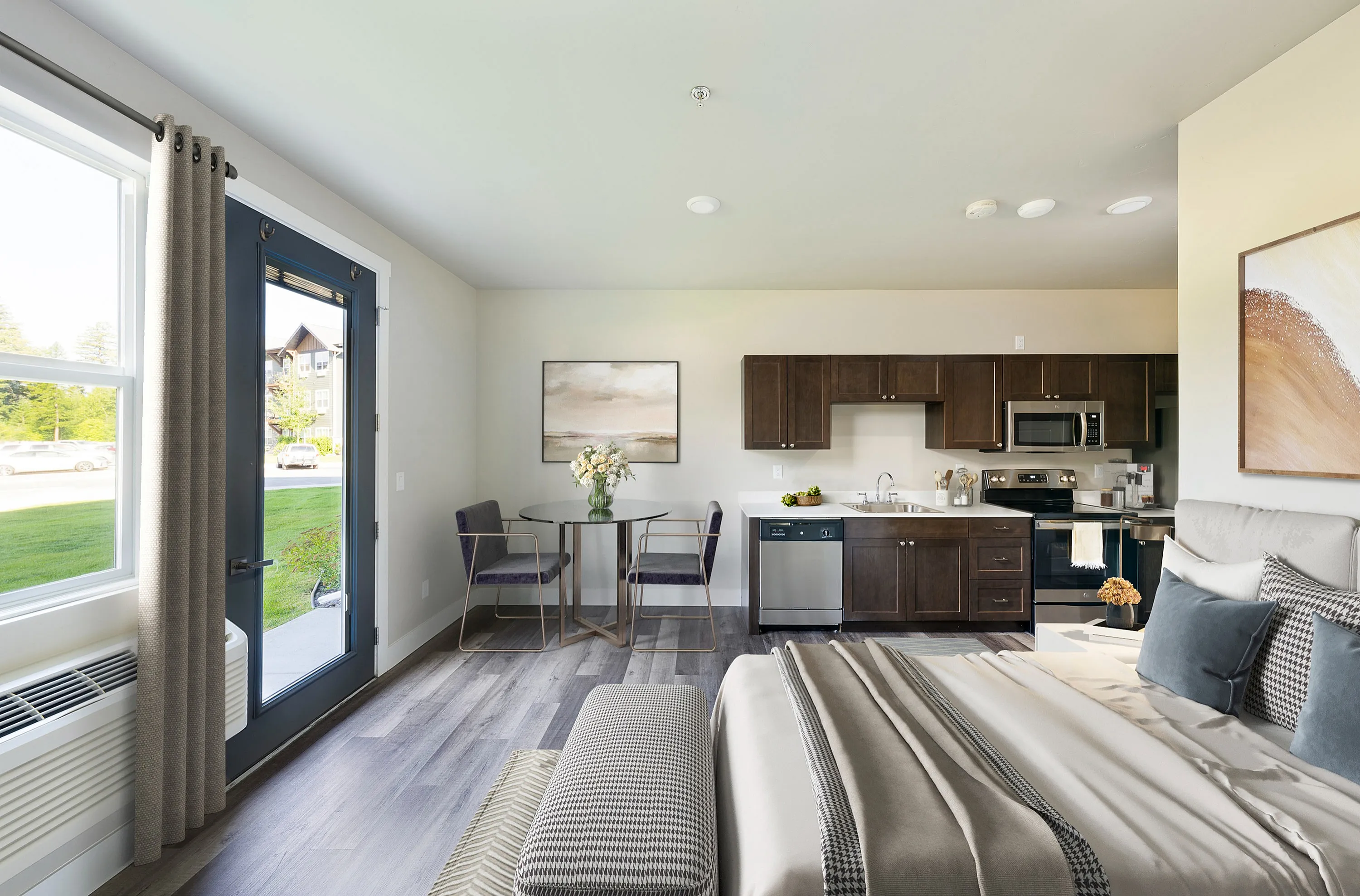 Bright and modern studio apartment featuring an open kitchen with dark wood cabinets, a cozy dining area with stylish chairs, and a comfortable bed with elegant bedding beside large windows overlooking greenery.