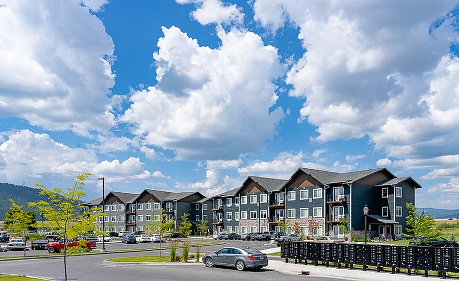Modern apartment complex on a sunny day with a vibrant landscape, ample parking, and picturesque blue skies with fluffy clouds.