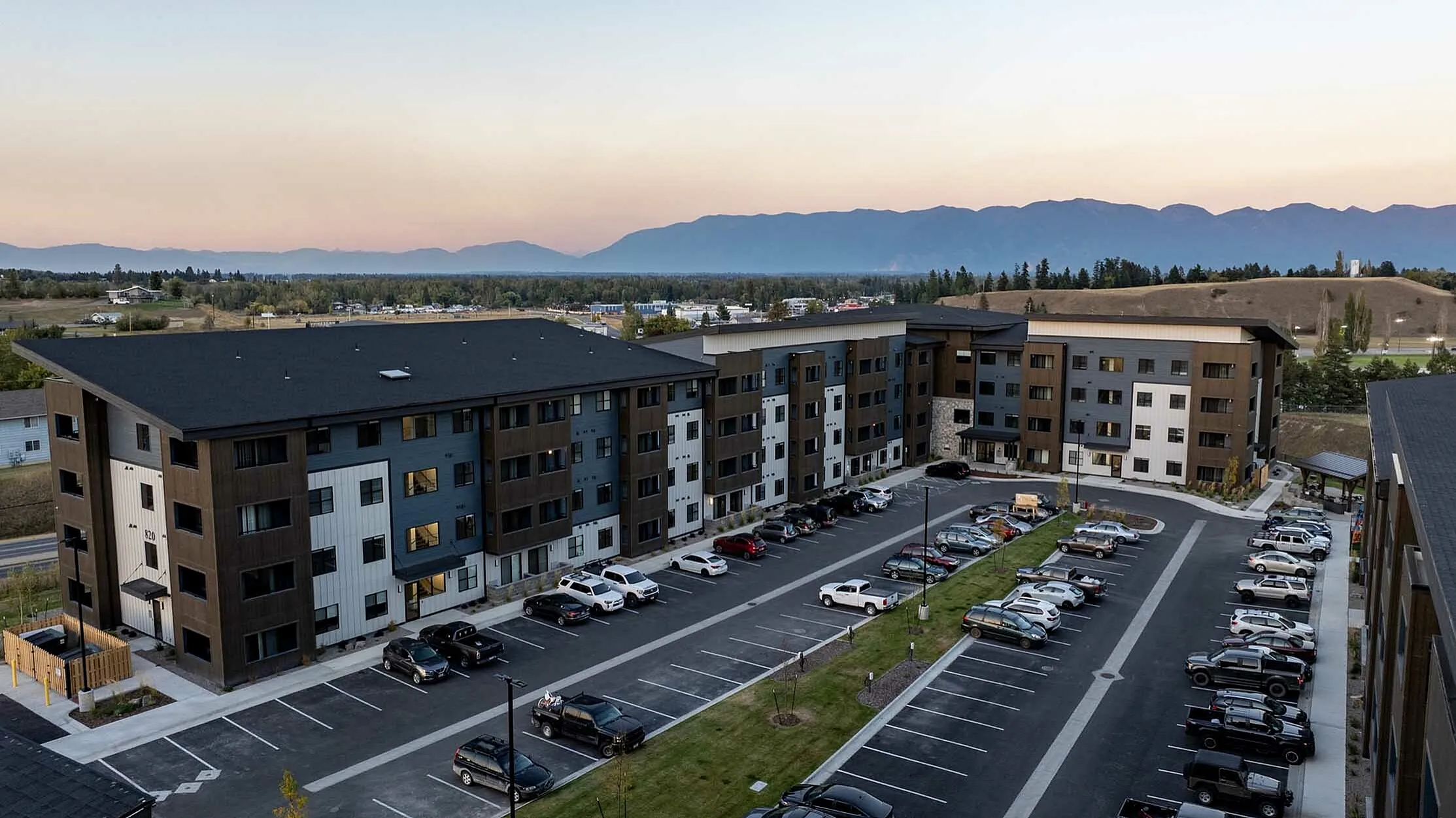 Modern apartment complex surrounded by scenic mountains at sunset, featuring ample parking and stylish architecture.
